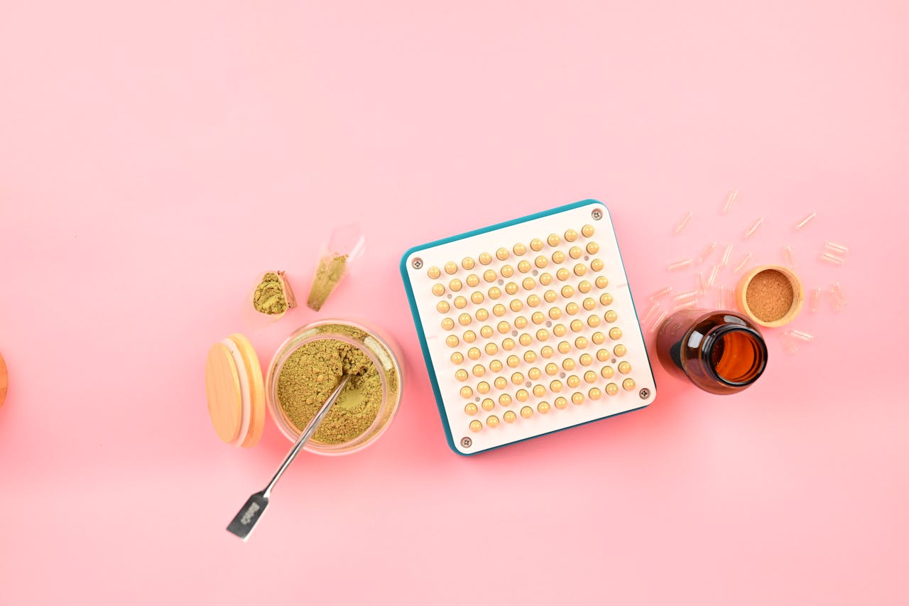 A capsule filling machine and herbs for supplement creation on a pink background.